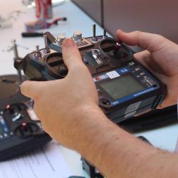 A person holds a remote controller, with another controller and a robot visible on a table behind.