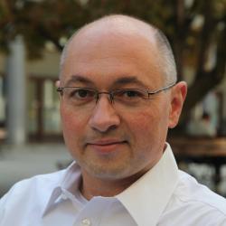 Markus Kraft head and shoulders in a white open button shirt and glasses with outdoor street backdrop.